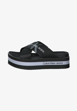 Czarna sandałka na platformie z krzyżującymi się paskami i białym napisem „Calvin Klein Jeans” na paskach i krawędzi podeszwy.