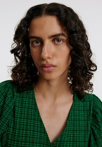 Groene jurk met patroon, een V-hals en geplooide mouwen, gemaakt van een gestructureerde stof en met een geruit ontwerp. Huidskleur is gemiddeld.