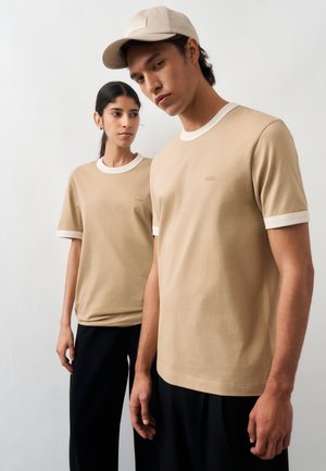 To modeller bærer beige T-shirts med hvid kant og sorte bukser; en bærer en beige kasket og poserer mod en simpel lys baggrund.