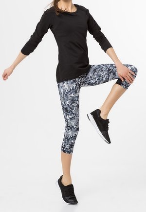 Haut de sport noir à manches longues, leggings capri imprimés avec un motif bleu et blanc, et chaussures de sport noires dans une pose de fitness.