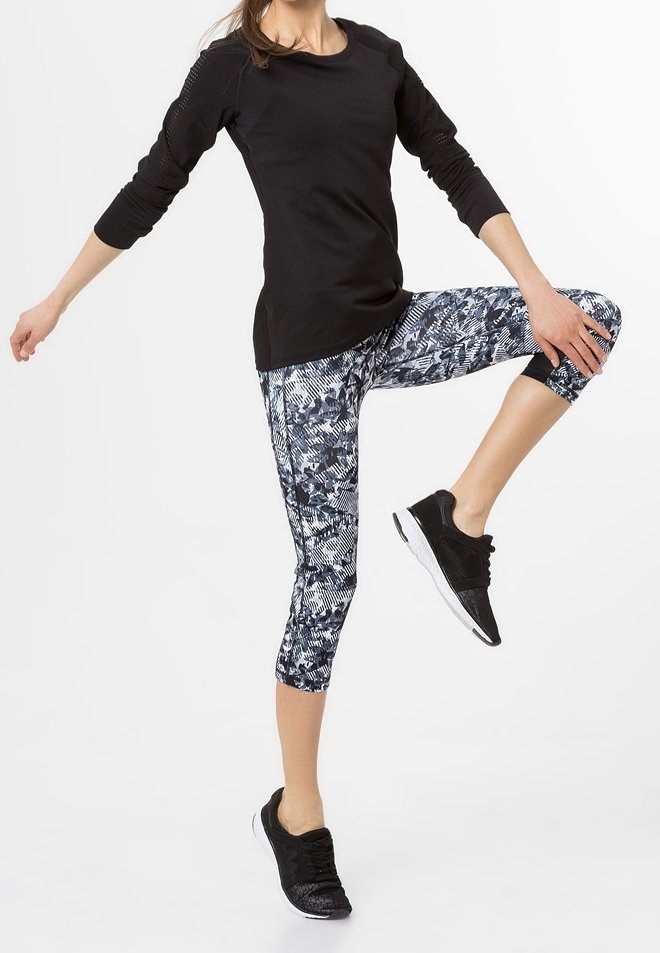 Haut de sport noir à manches longues, leggings capri imprimés avec un motif bleu et blanc, et chaussures de sport noires dans une pose de fitness.