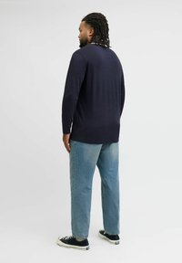 Vīrietis ar dreads frizūru, tērpies tumši zilos garos piedurkņu krekls, gaiši zili džinsi un melnām sporta kurpēm, stāv sānis uz balta fona.