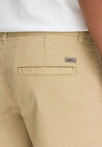 Beige Baumwollhosen mit einer strukturierten Oberfläche, ausgestattet mit einer Gesäßtasche und einem eingenähten Etikett mit der Aufschrift "Dedicated to Denim Perfection."