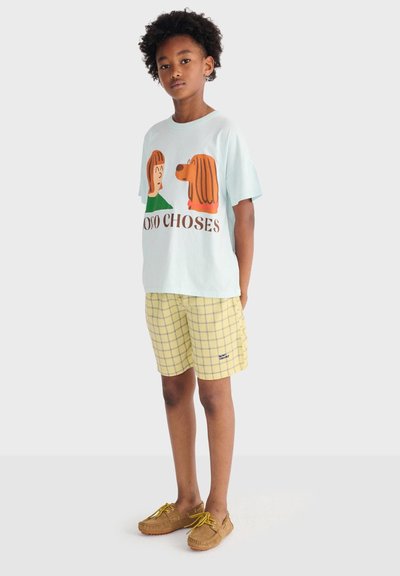 Enfant portant un t-shirt bleu clair avec une illustration de chien et de personne, un short à carreaux jaunes et des mocassins marron, debout devant un fond uni.