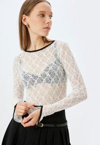 Witte kanten top met lange mouwen en zwarte accenten, met een bloemenpatroon en klokmouwen. Het model draagt het met een zwarte geplooide rok.