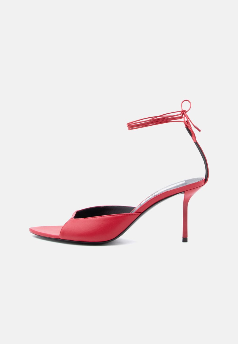 Κόκκινο σανδάλι με ψηλό τακούνι, μυτερή μύτη, λεπτά λουράκια στον αστράγαλο δεμένα σε φιόγκο και λεπτό τακούνι στιλέτο, εμφανιζόμενο από το πλάι σε λευκό φόντο.