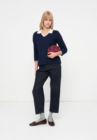 Pulover tricotat bleumarin cu decolteu în V și guler alb, purtat cu pantaloni din denim închis, șosete gri și balerini strălucitori burgundii. Ținând o geantă burgundie.