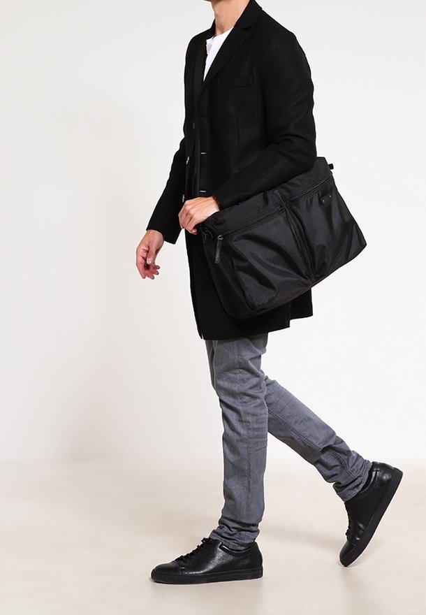 Homme marchant portant un manteau noir, un pantalon gris, des chaussures noires et portant un grand sac à bandoulière noir zippé.