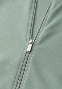 Close-up of a silver metal zipper on light green fabric, showing zipper teeth and pull tab in mid-zip position.