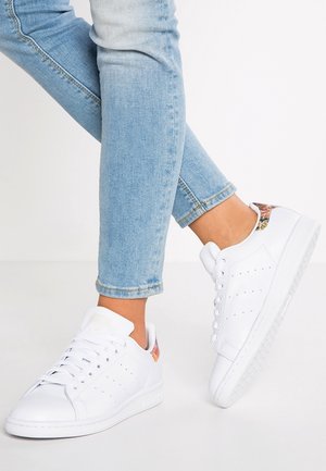 Person iført lyseblå jeans og hvide lave sneakers med blomsterdetalje på hælen, stående med den ene fod let løftet.
