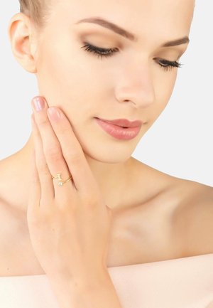 Gold ring with a delicate design featuring a small crystal accent. Model gracefully poses, holding her face with one hand. Smooth skin texture.