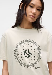 Beżowy bawełniany t-shirt z krótkim rękawem, z okrągłym wzorem graficznym w ciemnozielonym kolorze, z logo "KL" oraz szczegółami tekstowymi w pogrubionej czcionce.