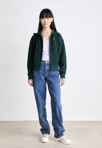 Grønn jakke med glidelås og lommer foran, brukt over en lys lilla t-skjorte. Kombinert med høyhalsede blå jeans og off-white sneakers.