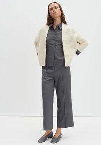 Donna che indossa un cardigan chunky color crema sopra una camicia grigia con bottoni, pantaloni grigi a gamba larga e scarpe grigie con tacco basso, su uno sfondo semplice.