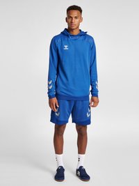 Blå idrottshoodie med texturerade axlar, i kombination med blå shorts med silverdetaljer. Bärs med vita ankelsockor och blå sneakers.