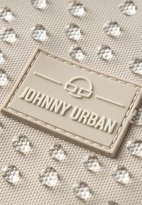 Tessuto beige con motivo a punti in rilievo e un'etichetta rettangolare di colore crema embossata con la scritta "JOHNNY URBAN." Dettaglio del bordo cucito visibile.