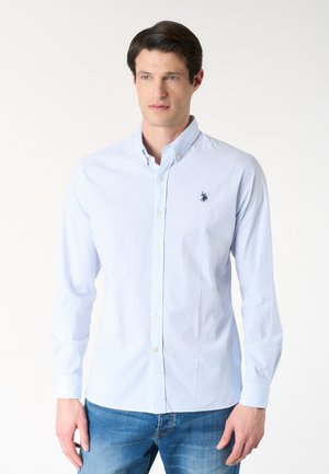 Uomo che indossa una camicia azzurro chiaro a maniche lunghe con sottili righe bianche e jeans blu, in piedi su uno sfondo bianco uniforme.