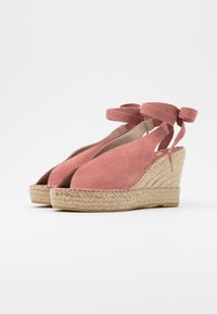 Růžové semišové sandály na klínu s propletenými juty podpatky a vázáním kolem kotníku, vystavené na bílém pozadí.