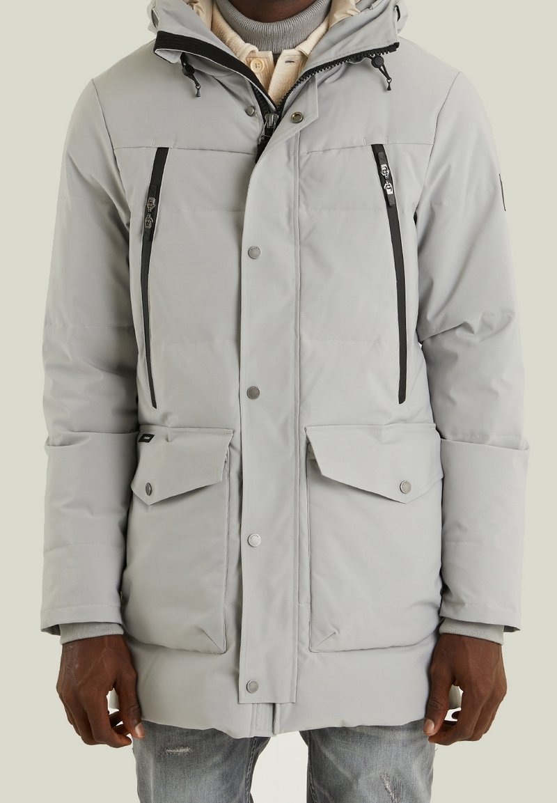 Homme portant un parka d'hiver gris clair avec des zips verticaux noirs sur la poitrine, des poches à rabat, et un tricot beige superposé en dessous.