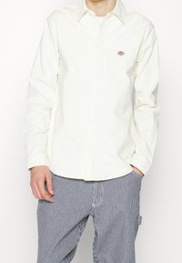 Camisa de manga larga color crema claro con cuello abotonado y pequeño logo bordado. Combina con pantalones oscuros de rayas finas.