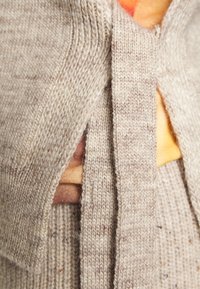 Gros plan sur un vêtement tricoté beige avec une ceinture nouée autour de la taille, montrant un tissu superposé et des détails de fil texturé.