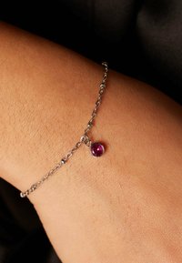 Zilveren kettingarmband met een ronde roze edelsteen hanger, tegen een huidgekleurde pols, met een gladde metallic textuur.