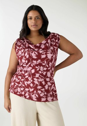 Donna con capelli scuri che indossa una canottiera bordeaux con motivo floreale rosa e pantaloni beige chiaro, posando con una mano sui fianchi.