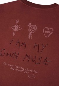 Tela de color rojo oscuro con texto bordado "SOY MI PROPIA MUSA" y diseños impresos de un ojo, una flor, un corazón con "ADORA" y una cita debajo.