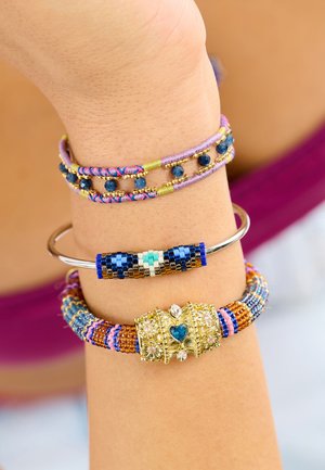 Trois bracelets au poignet : un bracelet de perles multicolores, un bracelet jonc en argent avec des perles bleues et blanches, et un bracelet manchette décoratif en or avec des pierres précieuses.