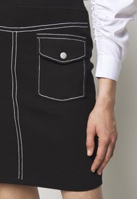 Schwarzer Strickrock mit weißer Naht, der eine Vordertasche mit Druckknopfverschluss aufweist. Der Stoff hat eine gerippte Textur. Leicht taillierte Passform.