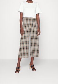 Bruine en beige geblokte wijde broek, gecombineerd met een witte t-shirt met ruches aan de mouwen en zwarte hoge sandalen.