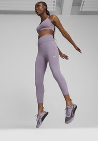 Šviesiai violetinė sportinė liemenėlė ir leggingai, pagaminti iš tampraus medžiagos, su aukšta juosmens juosta ir balta logotipu. Batai turi tinklelio dizainą su juodais akcentais.