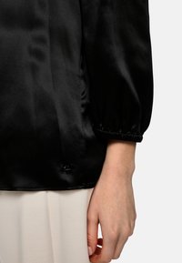 Black satin top with a relaxed fit, puffed sleeves, and gathered cuffs. Features a small logo detail near the hem.