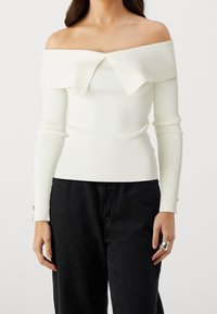 Weißes, schulterfreies, geripptes Oberteil mit weitem Kragen, langen Ärmeln und geknöpften Manschetten, kombiniert mit schwarzer Jeans.