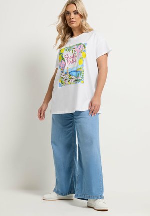 Vrouw met lang blond haar die een wit grafisch "Sicily Italia" T-shirt draagt, lichtblauwe jeans met wijde pijpen en witte sneakers, staand tegen een witte muur.