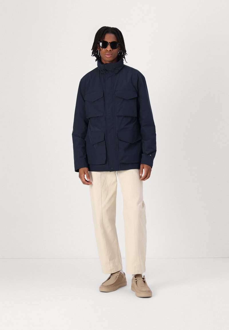 Navy isolierte Jacke mit hohem Kragen, mehreren Fronttaschen und einem Reißverschluss, kombiniert mit cremefarbenen Hosen und hellbraunen Schuhen.