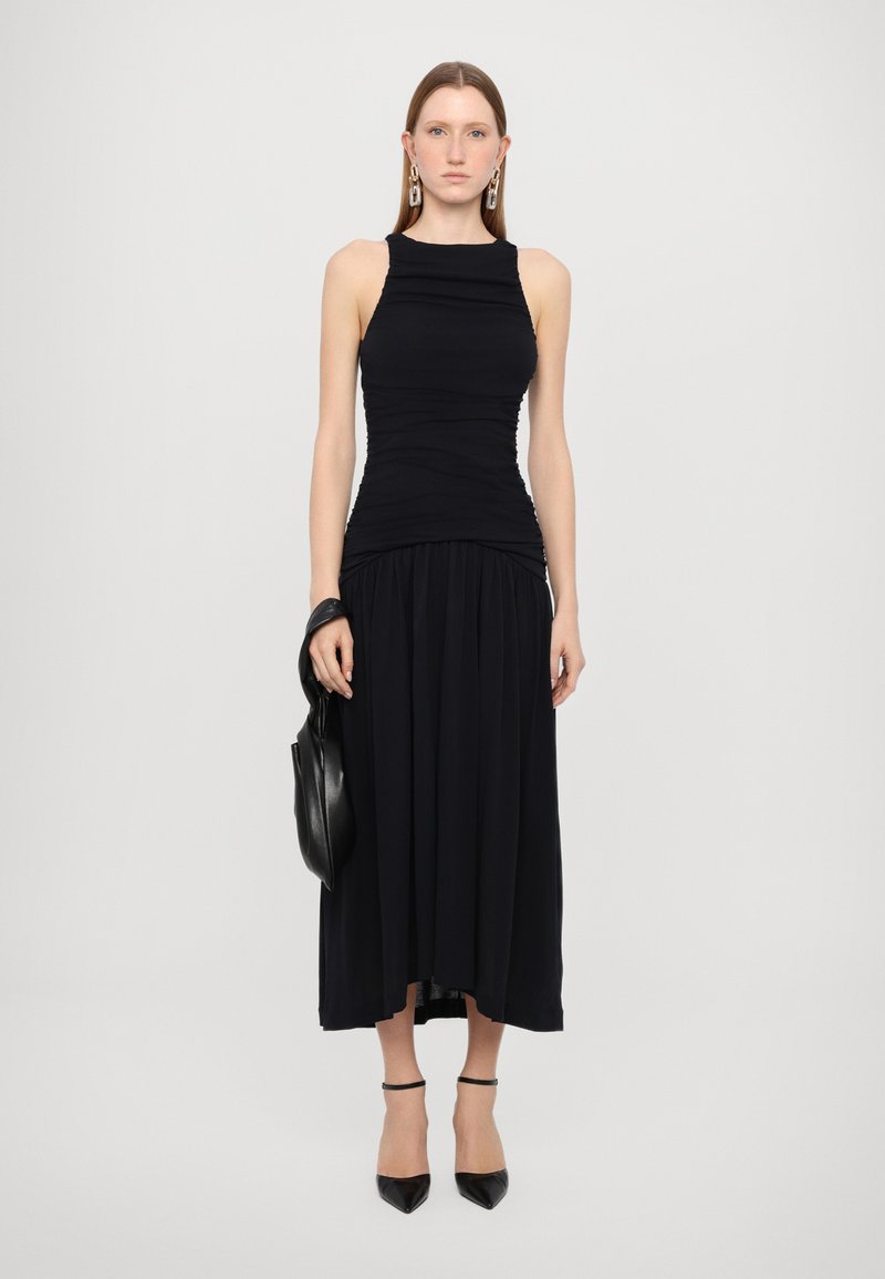 Schwarzes Kleid mit gerafftem Oberteil, ärmellosem Design und einem fließenden, plissierten Rock. Kombiniert mit schwarzen spitz zulaufenden Absätzen und einer eleganten Tasche.