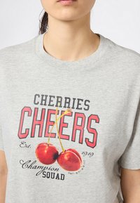 Osoba ubrana w jasnoszary t-shirt z czerwonymi wiśniami i napisem "Cherries Cheers Champion Squad Est. 1919" na przodzie.