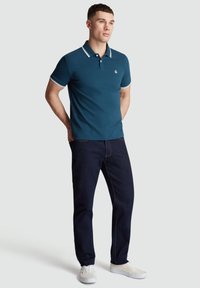 Donker teal polo shirt met contrasterende witte kraag en mouwtrim, gecombineerd met donkerblauwe jeans en witte sneakers.