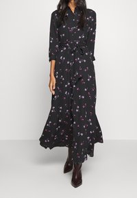 Zwarte jurk met lange mouwen en bloemenpatronen in roze en paars. Voorzien van een knopenlijn aan de voorkant, tailleband en een vloeiende silhouet.