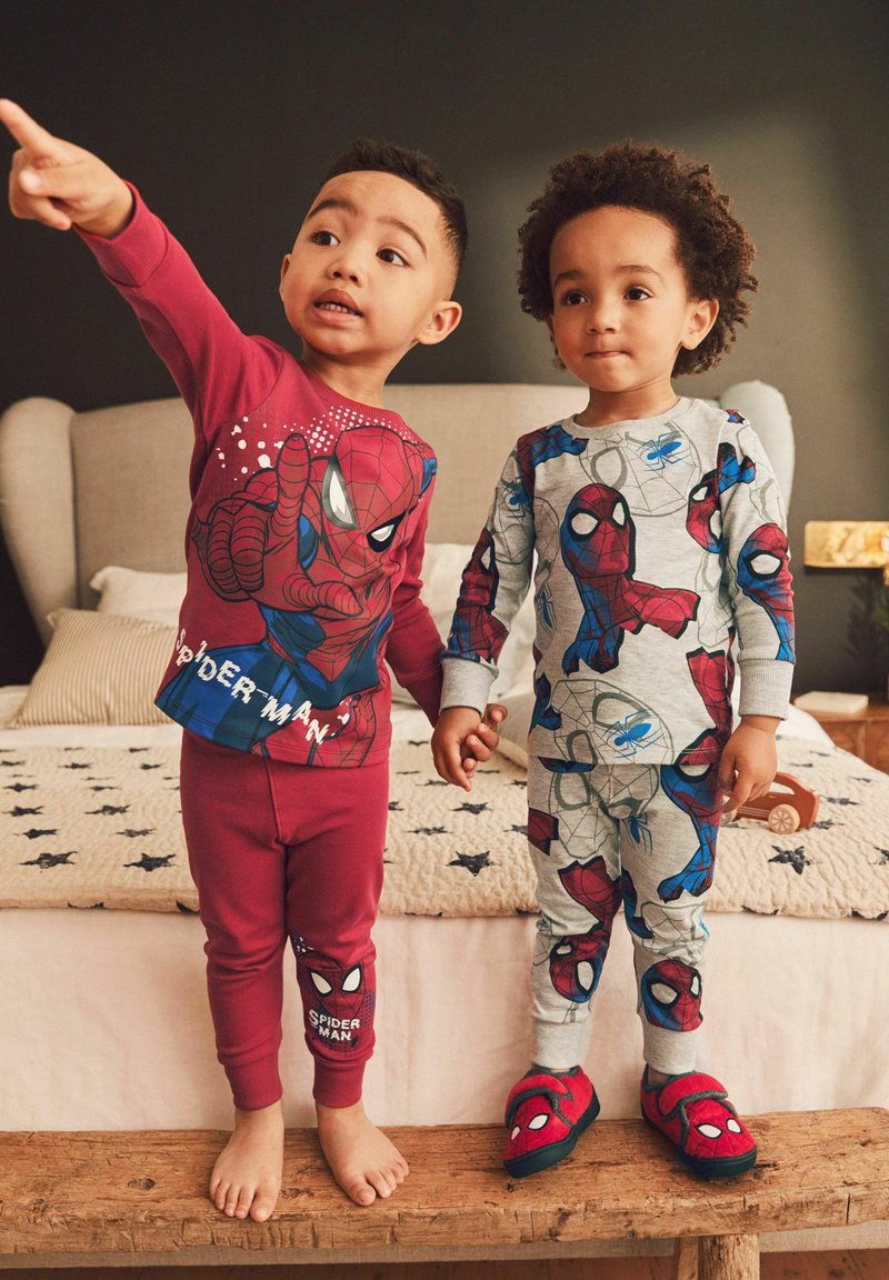 Zwei Kinder tragen Spiderman-Themen-Pyjamas. Das Kind links hat ein rotes Oberteil mit Grafik und passende rote Hosen. Das Kind rechts trägt Grau mit bunten Mustern und blauen Akzenten. Beide haben spielerisches Schuhwerk.
