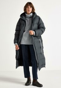 Nainen seisoo päällään pitkä musta untuvatakki, harmaan pooloneuleen, tummansiniset farkut ja mustat nilkkurit valkoista taustaa vasten.