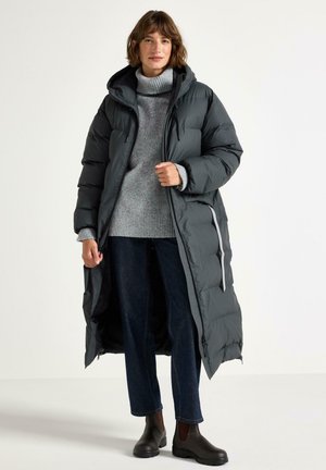 Kvinde stående iført en lang sort dunjakke over en grå turtleneck sweater, mørkeblå jeans og sorte ankelstøvler mod en hvid baggrund.