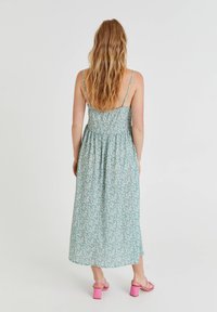 Femme aux longs cheveux ondulés portant une robe midi verte à fleurs avec de fines bretelles et des sandales à talons blocs roses, debout de dos.