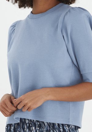 Femme portant un haut bleu clair à manches courtes avec des épaules bouffantes et une jupe noire et blanche à motifs, les mains ajustant l'ourlet du haut.