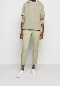 Sudadera de un suave color verde oliva claro con un bolsillo canguro en la parte frontal, combinada con chinos entallados a juego y zapatillas blancas. Material de algodón suave.