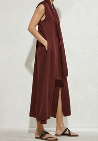 Ärmelloses, bodenlanges burgunderrotes Kleid mit einem Schlitz vorne und einer Einschubtasche, getragen mit schwarzen flachen Sandalen auf hellem, strukturiertem Hintergrund.