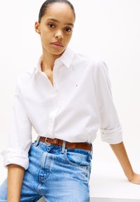 Camisa blanca de botones con mangas remangadas, combinada con jeans de denim azul claro de tiro alto y un cinturón marrón. Detalle de logo sencillo en la camisa.