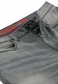 Jeans de denim gris con cremallera, cierre de botón y una cintura con logo rojo. La tela presenta un efecto desgastado y desteñido con ligera texturización.