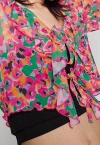 Blouse à volants floraux vifs avec des motifs roses, verts, oranges et violets noués en nœuds sur un haut court noir, porté sur le torse d'une personne.